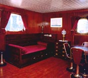 Part of a combined living- and bedroom onboard a ship, furnished with restored 20th century ship's bunk bed sofa, chest of drawers, table, swivel chairs and stool, as well as original kerosene lamps.