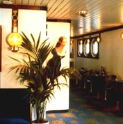 Part of an entrance hall, onboard a hotel ship, furnished with restored 20th century ship's tables and swivel chairs, decorated with figurehead and equipped with newlymade brass lamps. 