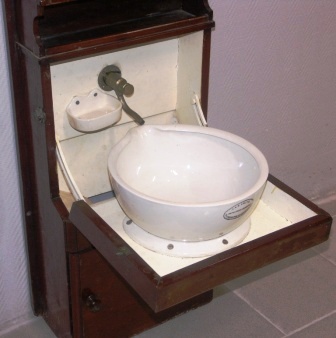 Mahogany wash cabinet for wall mounting with porcelain basin and brass tap. Made by A & R Smith Glasgow. Incl mirror, shelf as well as holder for glasses and decanter. Water container and portable wastewater bucket missing.