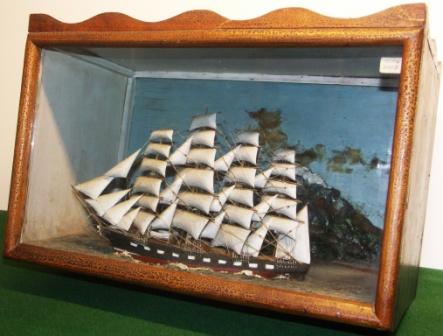 19th century sailor-made diorama depicting a four-masted barque with set sails. Decorative background with mountainous coastline, village and lighthouse. 