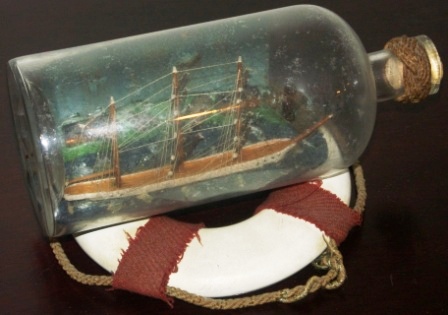 Late 19th century sailor-made ship model housed in bottle depicting a 3-masted barque. Incl lifebuoy-stand.